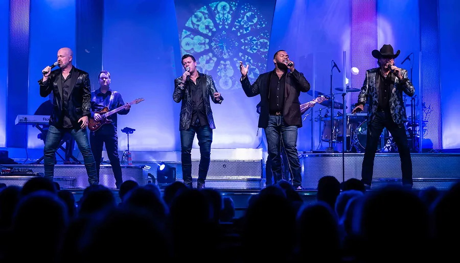 Four male vocalists are performing on stage with a live band and a vibrant light display in the background, entertaining an audience.