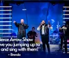 Four singers are energetically performing on stage with a band in the background illuminated by colorful stage lighting