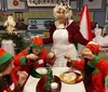 A person dressed as Mrs Claus looks surprised in a Christmas-themed kitchen while surrounded by individuals dressed as elves who are focused on preparing holiday treats