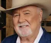 The image shows a smiling man with a white mustache and beard wearing a cowboy hat and a blue blazer with a rustic wooden background