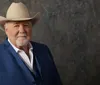 The image shows a smiling man with a white mustache and beard wearing a cowboy hat and a blue blazer with a rustic wooden background