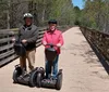 Two people are standing with Segways on a wooden bridge outdoors wearing helmets and smiling for the camera