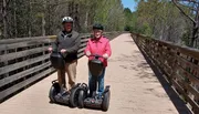 Two people are standing with Segways on a wooden bridge outdoors, wearing helmets and smiling for the camera.
