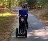 Two people are standing with Segways on a wooden bridge outdoors wearing helmets and smiling for the camera