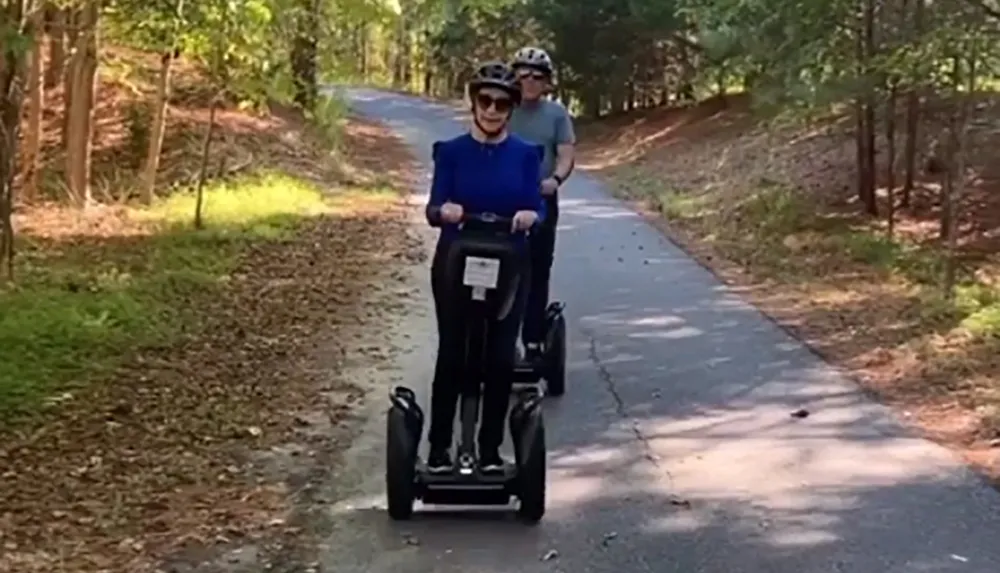 Two individuals are riding Segways on a paved path through a wooded area