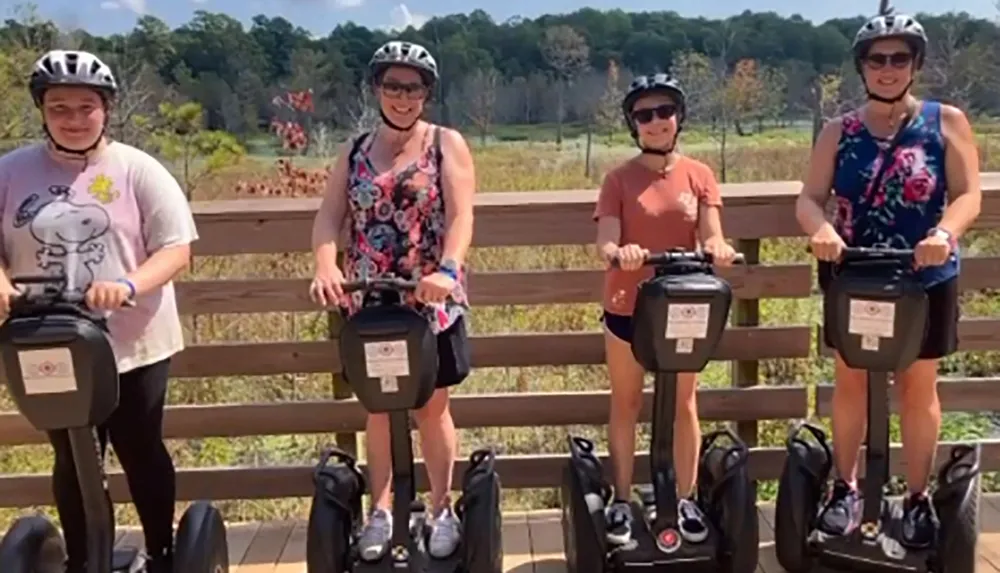 Four individuals wearing helmets are poised for a group photo while standing with their personal transporters on a wooden deck in a sunny outdoor setting