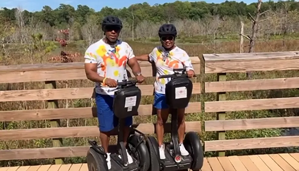 Two individuals wearing helmets and matching shirts are standing on Segways on a wooden boardwalk outdoors
