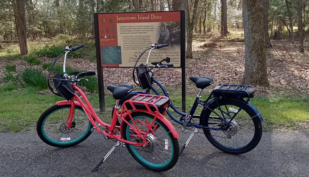 Two bicycles one red and one blue are parked in front of an informational sign about Jamestown Island Drive surrounded by a wooded area