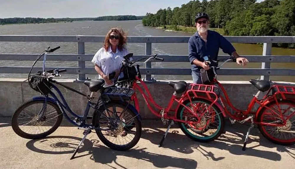 Two people are standing with their bicycles on a bridge with a lush riverscape in the background