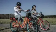 Two individuals wearing helmets are riding colorful bicycles across a grassy field.