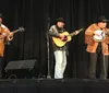 Three musicians playing a double bass guitar and banjo perform with visible enthusiasm in front of a wooden backdrop