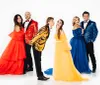 Five people wearing colorful formal attire pose in front of a white background with two couples on the sides and a man in the center playfully pouting towards a woman in yellow