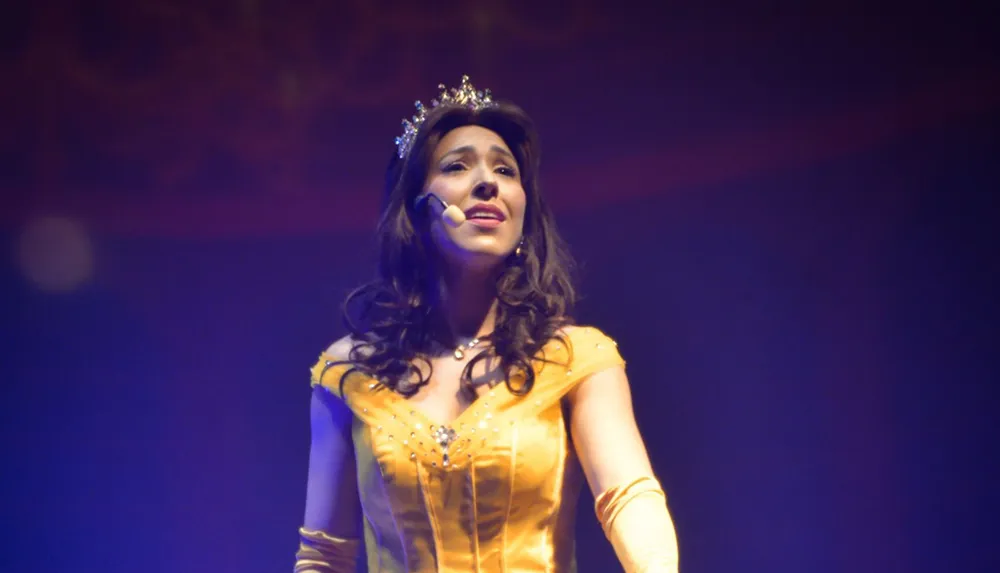 A woman dressed as a princess in a yellow gown and tiara is performing on stage with a microphone headset