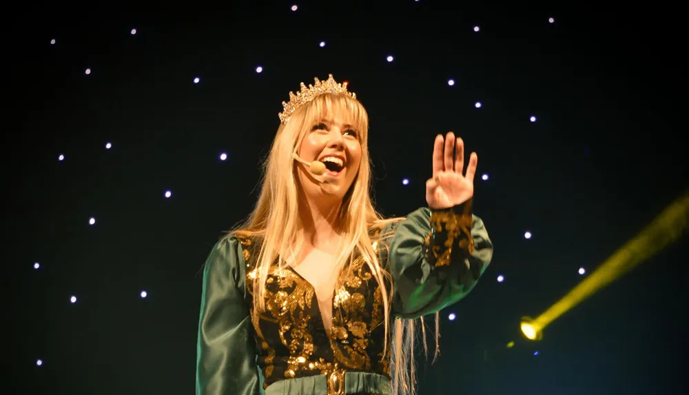 A woman wearing a crown and a microphone headset is performing on stage reaching out with her hand against a backdrop of shining lights