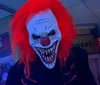 A person is wearing a scary clown mask with bright red hair and a wide menacing grin standing indoors under a blue light