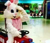 A person dressed in a large fluffy bunny costume is seated on a red childrens ride-on train in an indoor play area