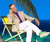 A person is smiling and relaxing on a beach chair next to a palm tree with a clear blue sky and sea in the background