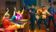 The image shows a vibrant and colorful scene with performers in retro-style attire, enthusiastically playing music, with one person lying on a piano playing keys upside down.