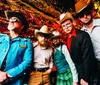 A group of five men sporting cowboy hats and eclectic outfits pose confidently with a festive colorful tinsel background accompanied by a quote praising their wit and musical talent attributed to the Los Angeles Times