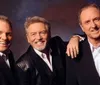 Three smiling men one holding a guitar pose together for a friendly portrait against a gray background