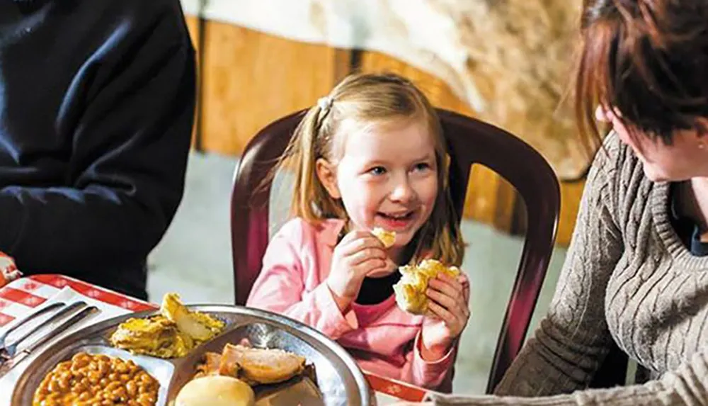 A smiling young girl is sitting at a table with a plate of food interacting with someone beside her