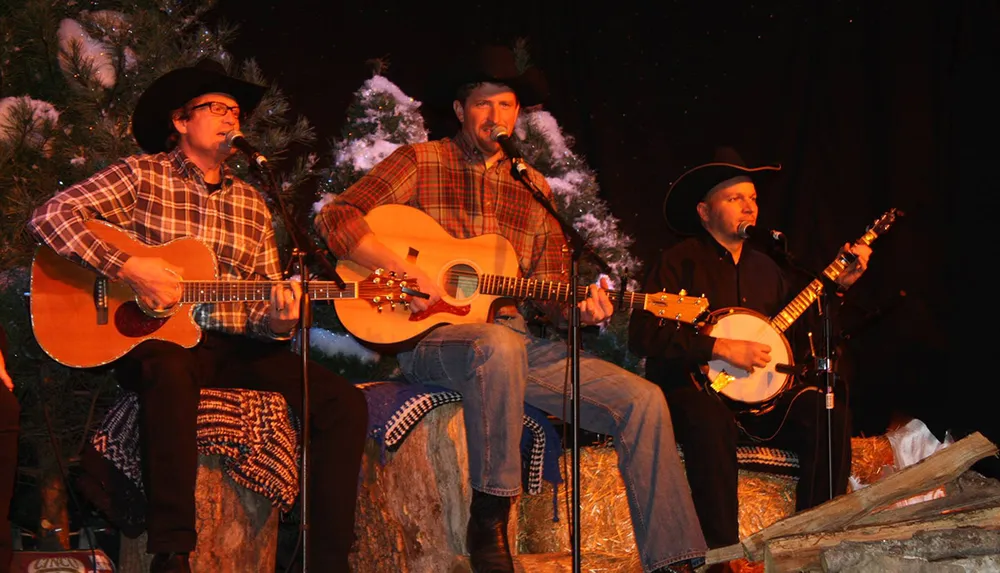 Three musicians in cowboy hats are performing with guitars and a banjo on a rustic stage with snowy trees in the background