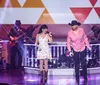 A performer is singing on stage while a group of dancers with cowboy hats performs a synchronized routine behind him all set against a vibrant light display