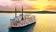 A paddle steamboat adorned with flags and bunting sails on a calm body of water during a vibrant sunset.
