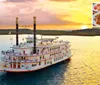A paddle steamboat adorned with flags and bunting sails on a calm body of water during a vibrant sunset