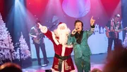 A performer in a sparkly suit is singing on stage alongside someone dressed as Santa Claus, with a festive backdrop and musicians playing in the background.