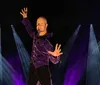 A person wearing a purple velvet shirt is posing dramatically with raised arms against a dark background with beams of blue and purple light