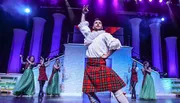 A performer in a kilt and period shirt energetically dances on stage while other dancers in traditional and formal attire clap along in the background, set against a colorful theatrical backdrop.