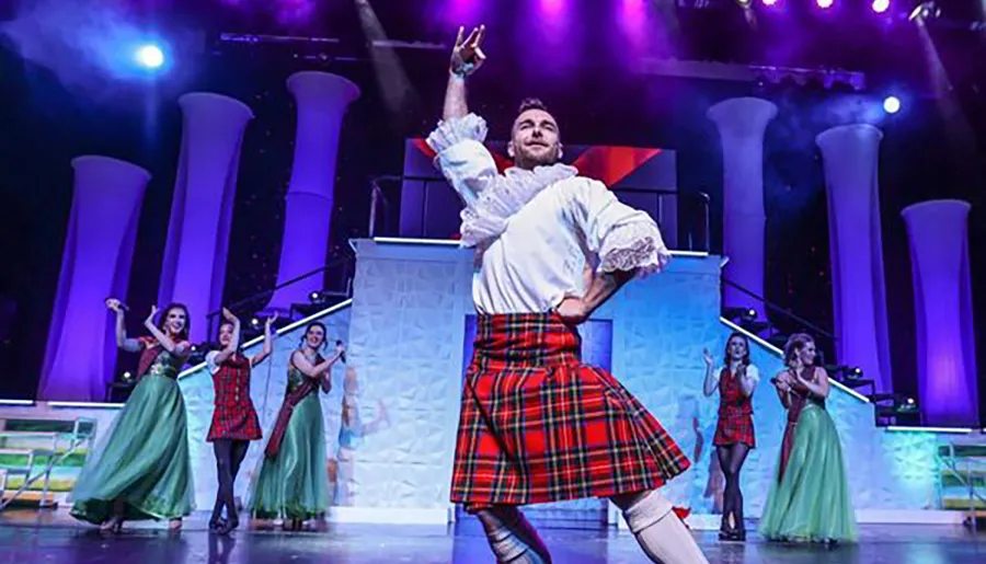 A performer in a kilt and period shirt energetically dances on stage while other dancers in traditional and formal attire clap along in the background, set against a colorful theatrical backdrop.