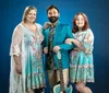 Three smiling people are posing together with the central figure holding a blue guitar and wearing a bright blue suit flanked by two women in patterned dresses on a white background