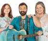 Three smiling people are posing together with the central figure holding a blue guitar and wearing a bright blue suit flanked by two women in patterned dresses on a white background