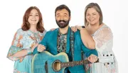 Three smiling people are posing together, with the central figure holding a blue guitar and wearing a bright blue suit, flanked by two women in patterned dresses on a white background.