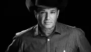 The image depicts a black and white portrait of a man wearing a cowboy hat and a patterned shirt, gazing directly at the camera against a black background.