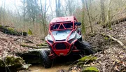A red off-road vehicle is navigating a rocky creek bed, with a person visible behind the roll bars, engaged in an outdoor adventure.