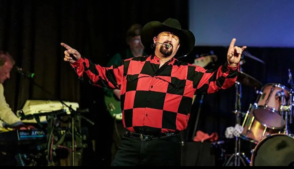 A person in a cowboy hat and red-and-black checked shirt is singing passionately with outstretched arms on stage while a band performs in the background