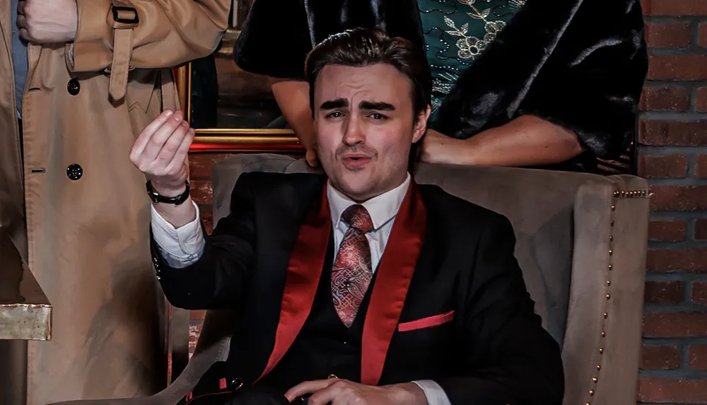A person in a suit with a red satin collar is making a expressive gesture while sitting in a chair