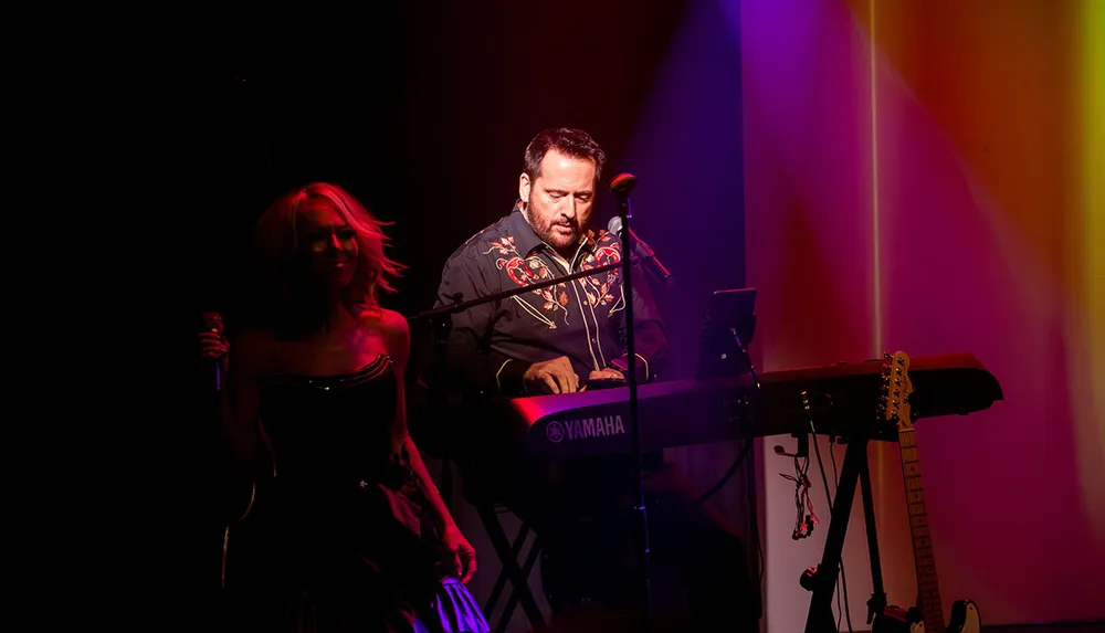 A musician is playing a Yamaha keyboard on stage under colorful lighting with a singer standing nearby holding a microphone