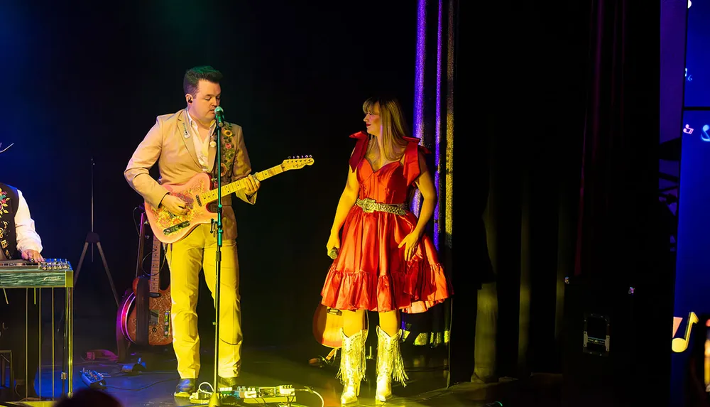 A man and a woman in vibrant costumes perform on stage with the man playing a guitar and the woman holding a microphone