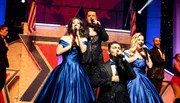 A group of performers in elegant blue gowns and sparkly suits sing into microphones on a brightly lit stage.