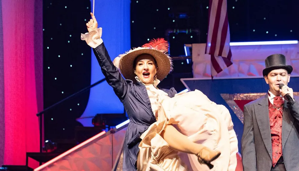 A performer in a vintage-style costume energetically kicks up her leg on stage beside a man in formal attire