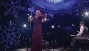 A woman is playing the violin on stage next to a man at a grand piano, with a backdrop of snowflake decorations and dramatic lighting.
