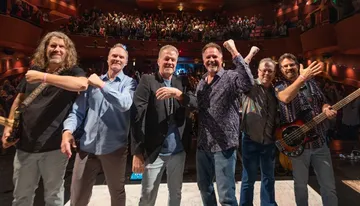 A group of six men poses and smiles on stage in front of an applauding audience in a theater.