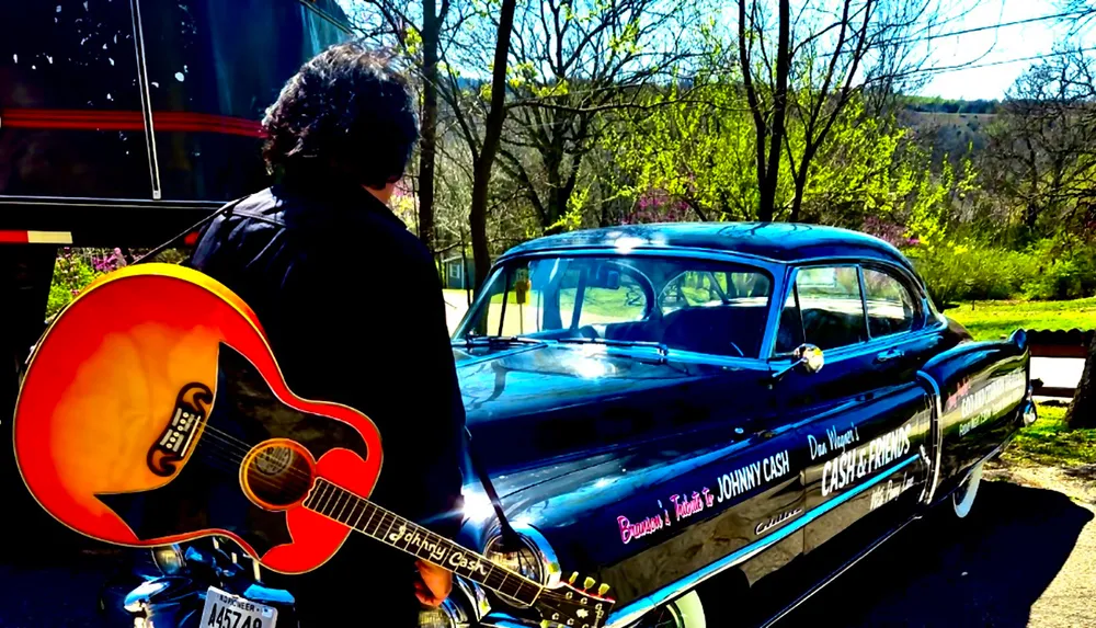 A person with a guitar stands in front of a vintage black car adorned with Johnny Cash tribute text parked outdoors