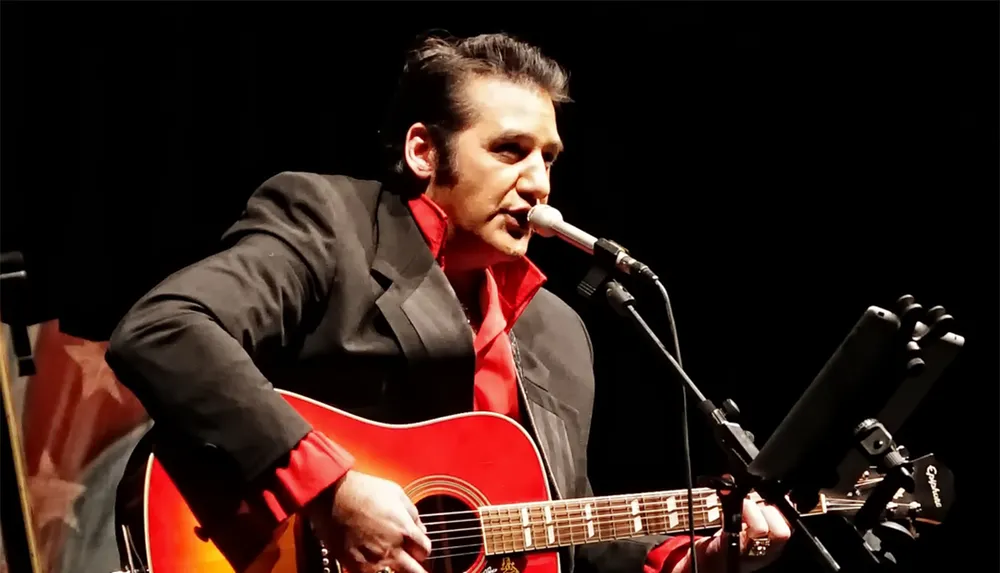 A musician in a suit plays an acoustic guitar and sings into a microphone on stage