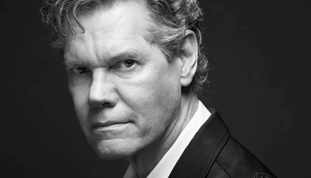 The image is a black and white close-up portrait of a man with curly hair wearing a suit and looking directly at the camera