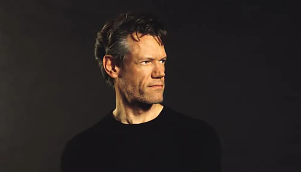 A man is gazing to the side against a dark background wearing a black shirt